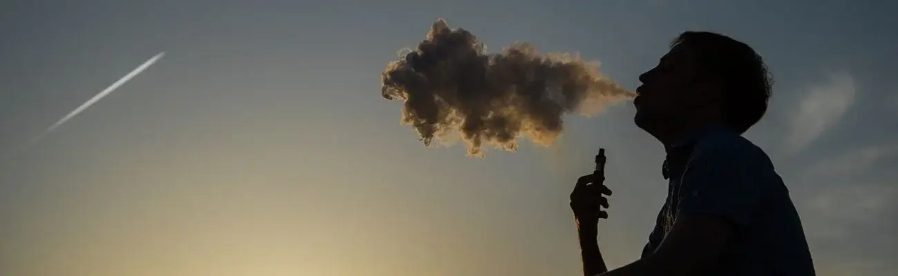 A man smoking a vape at night, blowing out a big cloud of vaporize smoke.